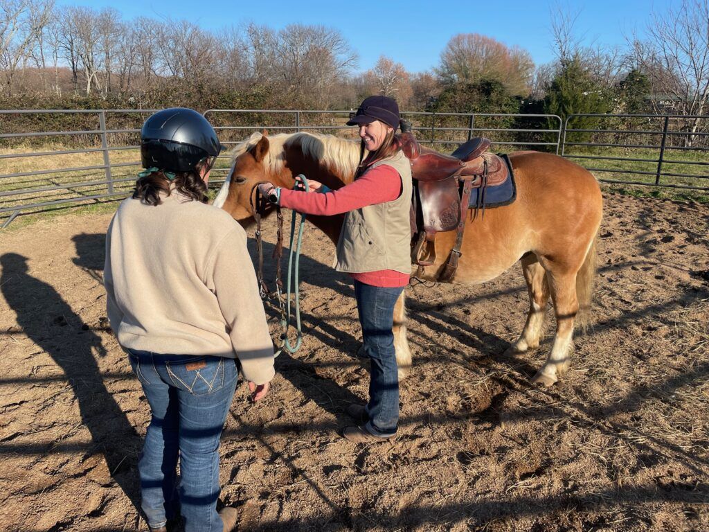 Woman wearing pink long sleeve shirt, blue jeans and beige vest demonstrates how to bridle a horse for an another adult female wearing a riding helmet, blue jeans and beige sweatshirt