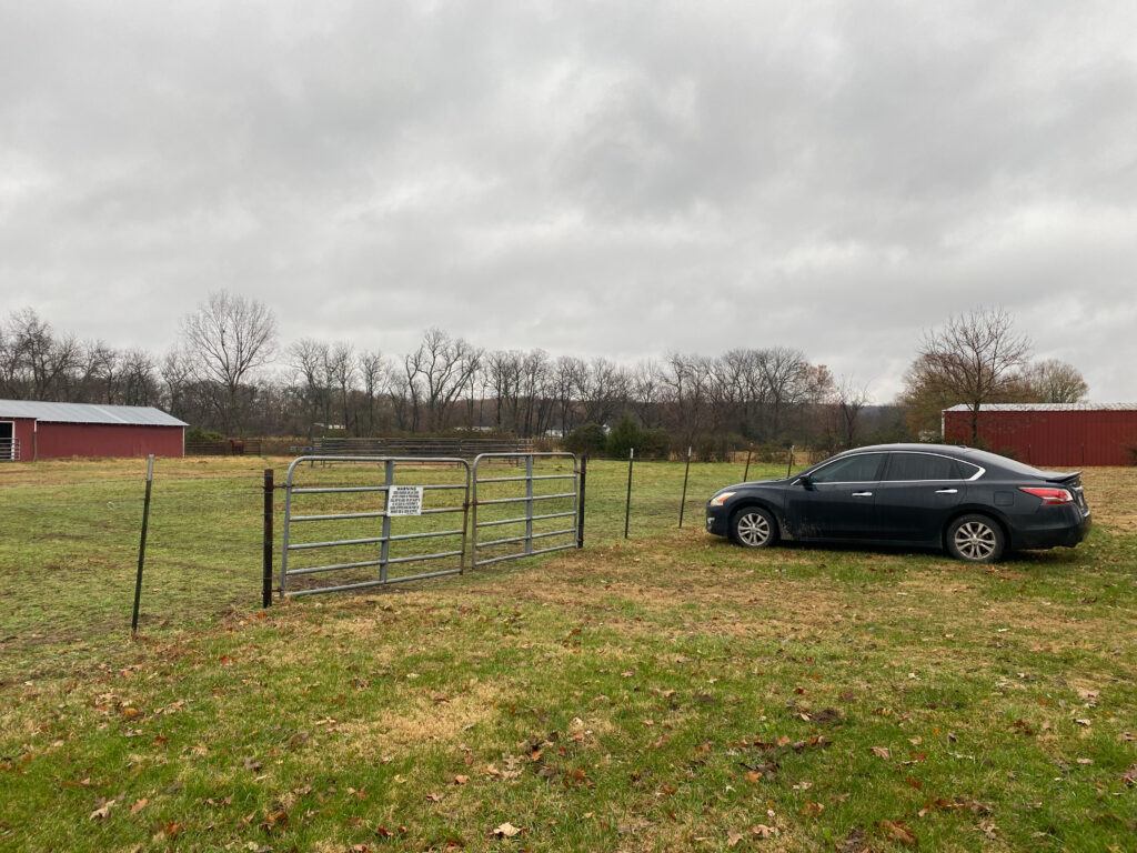 black sedan parked in front of a barbed wire fence bordering a pasture, sky is cloudy and overcase, trees are bare, there is a red barn in the background