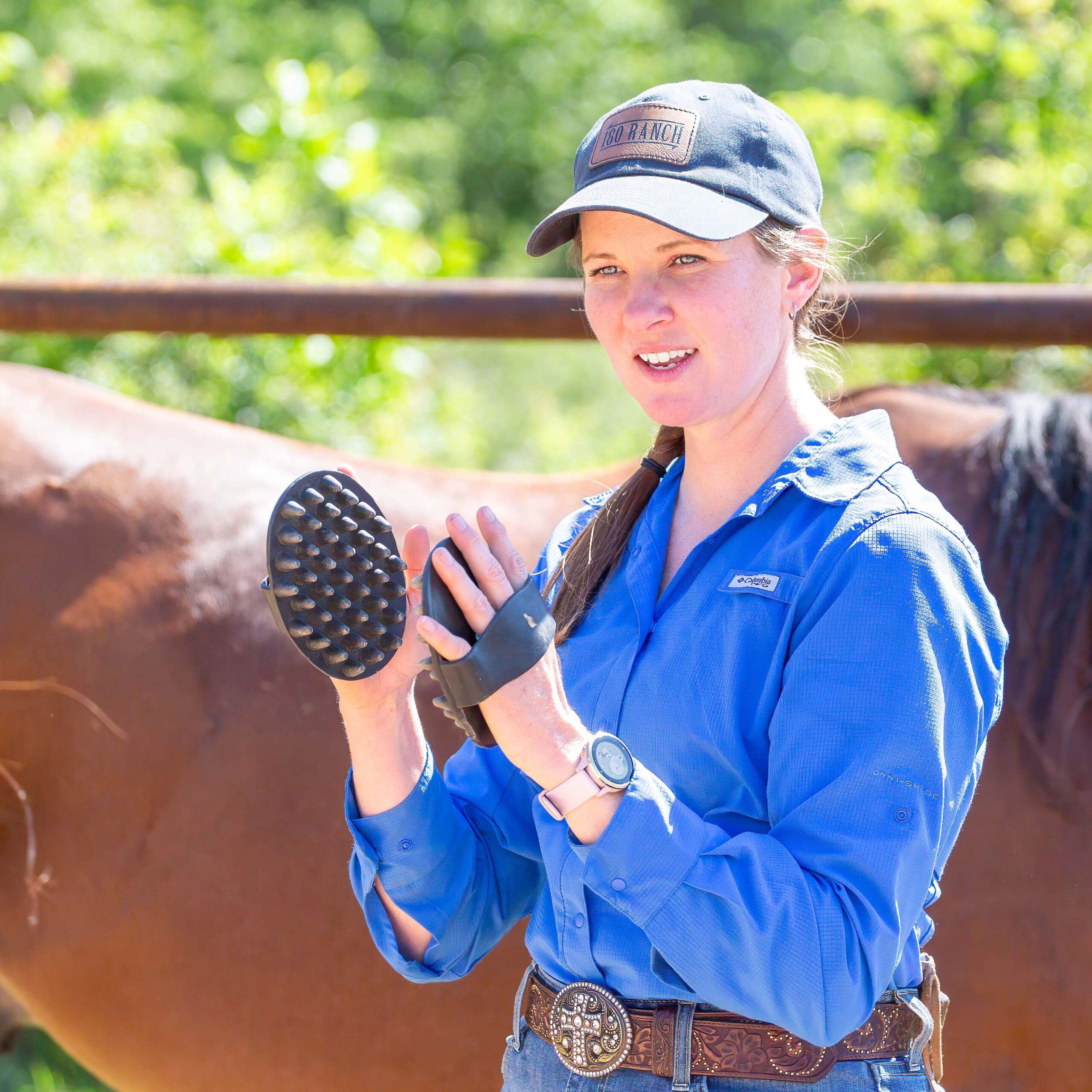 Woman in blue ball cap and blue collared button up shirt demonstrates how to groom a horse with rubber curry comb