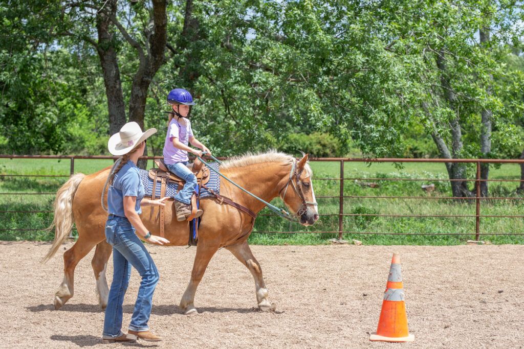 Woman in a straw cowboy hat, blue shirt and blue jeans teaches a young girl how to ride a horse. The girl is wearing a purple helmet and riding a golden horse with a blond mane and tail.