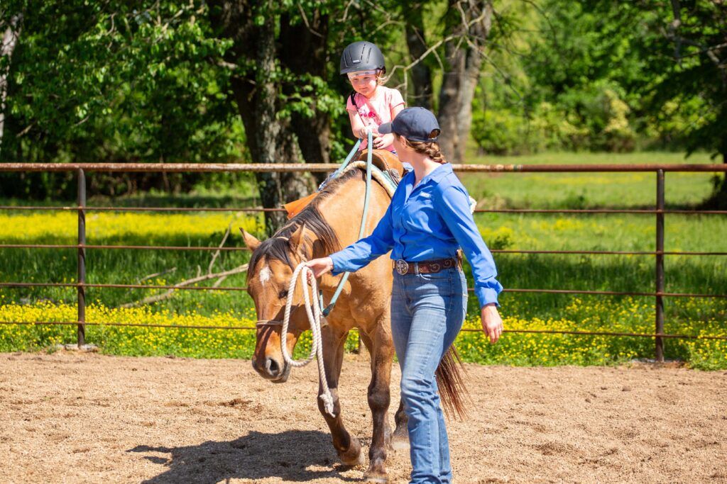 woman in blue shirt, jeans and blue ball cap leads a horse with a young girl wearing a pink shirt and riding helmet