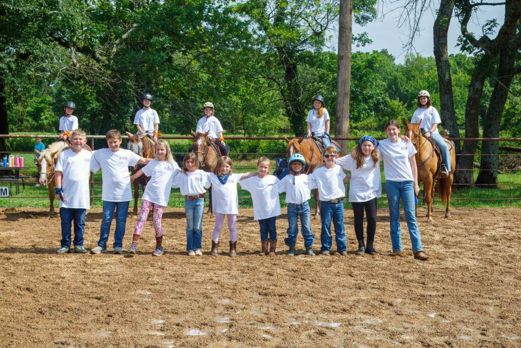 Group of smiling children at a horse camp in northwest Arkansas. Nine of the kids have their arms around each others' shoulders. There are five kids on horseback behind them.
