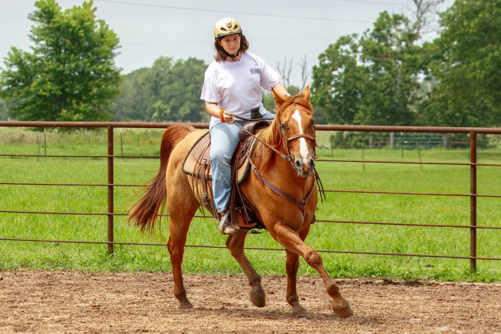 Teenage girl with short brown hair and a beige riding helmet rides a red dun horse in a western saddle.